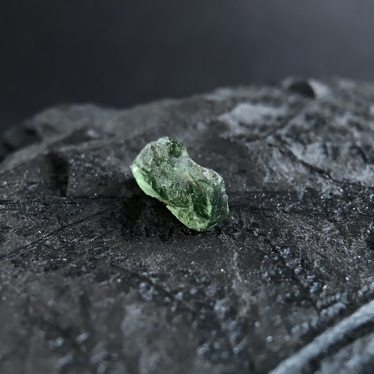 raw moldavite on a textured black surface with a dark background