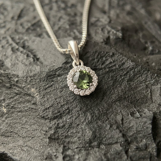 Silver pendant necklace with a round moldavite  haloed by small zirconia, resting on textured dark rock.