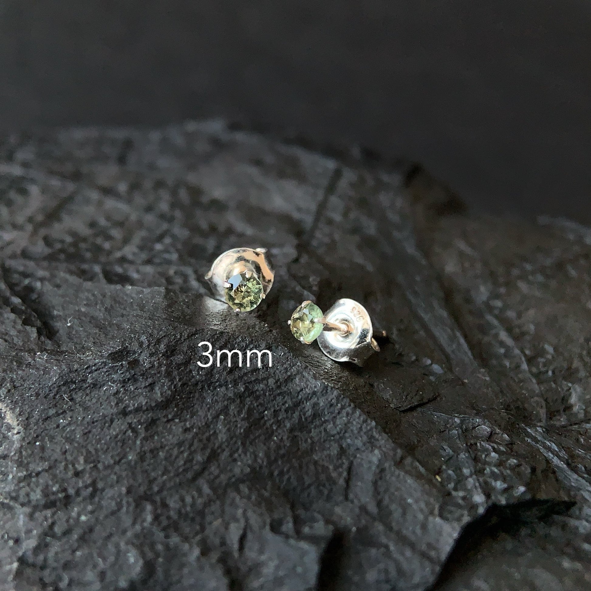 Small silver earrings with Moldavite on a textured dark surface, labeled '3mm'.