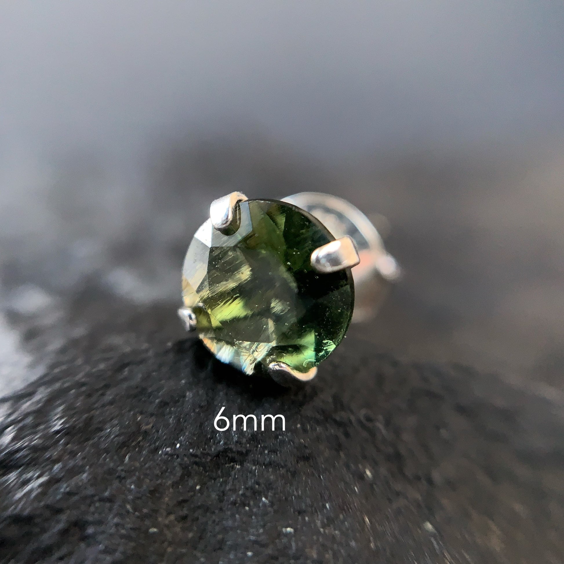 Macro shot of Moldavite stud earring on a dark textured surface with '6mm' text.