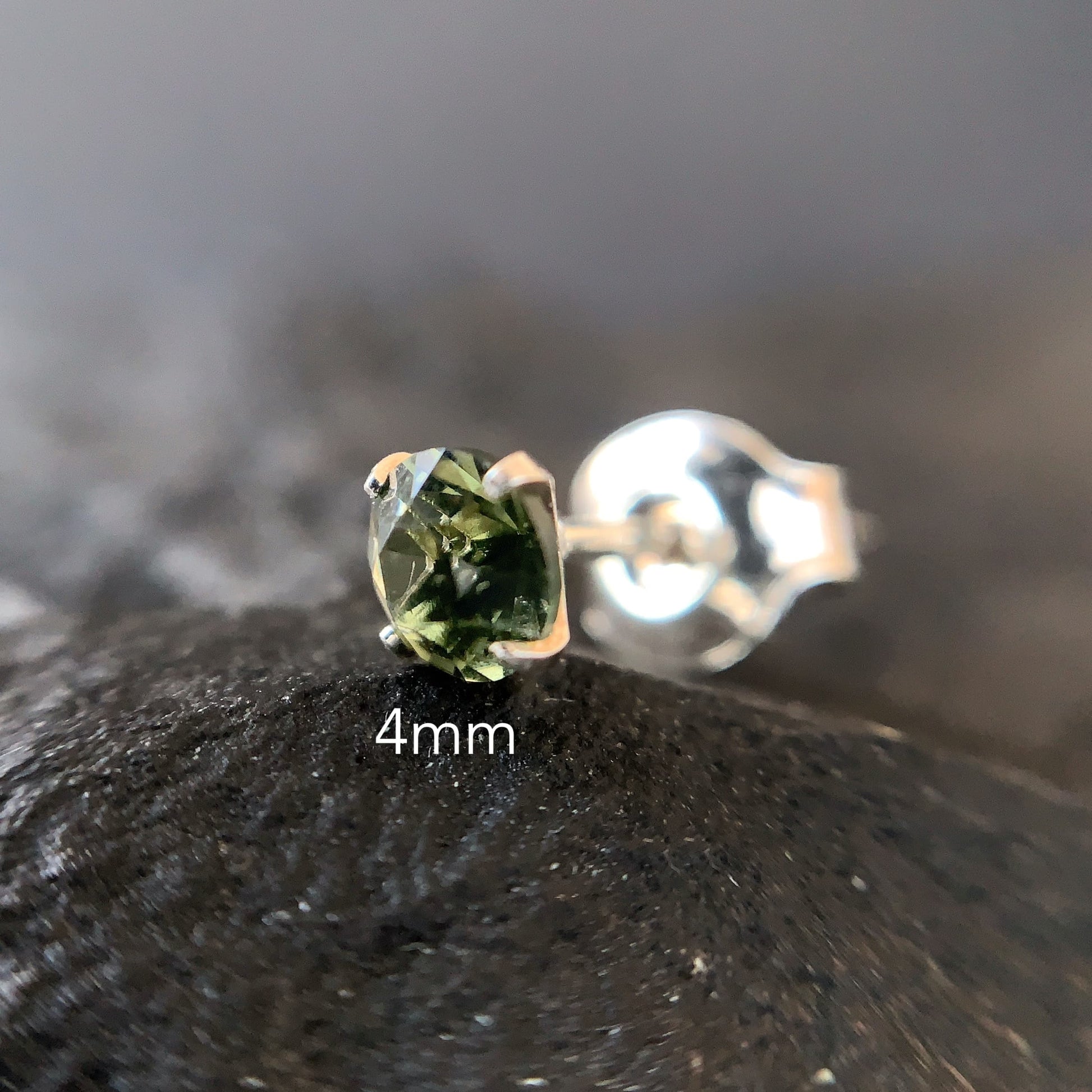 Close shot Silver stud earring with a 4mm moldavite on a dark textured surface.