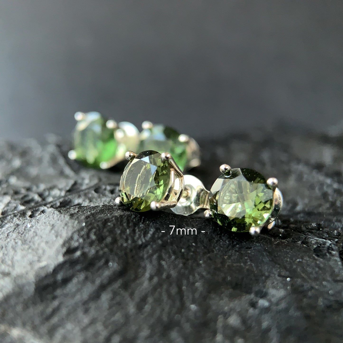 Moldavite stud earrings on a dark textured surface in 7 mm setting