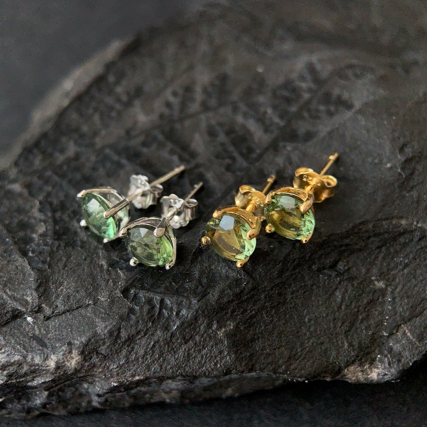 Pair of gold plated and silver stud earrings with green gemstones on a dark stone surface