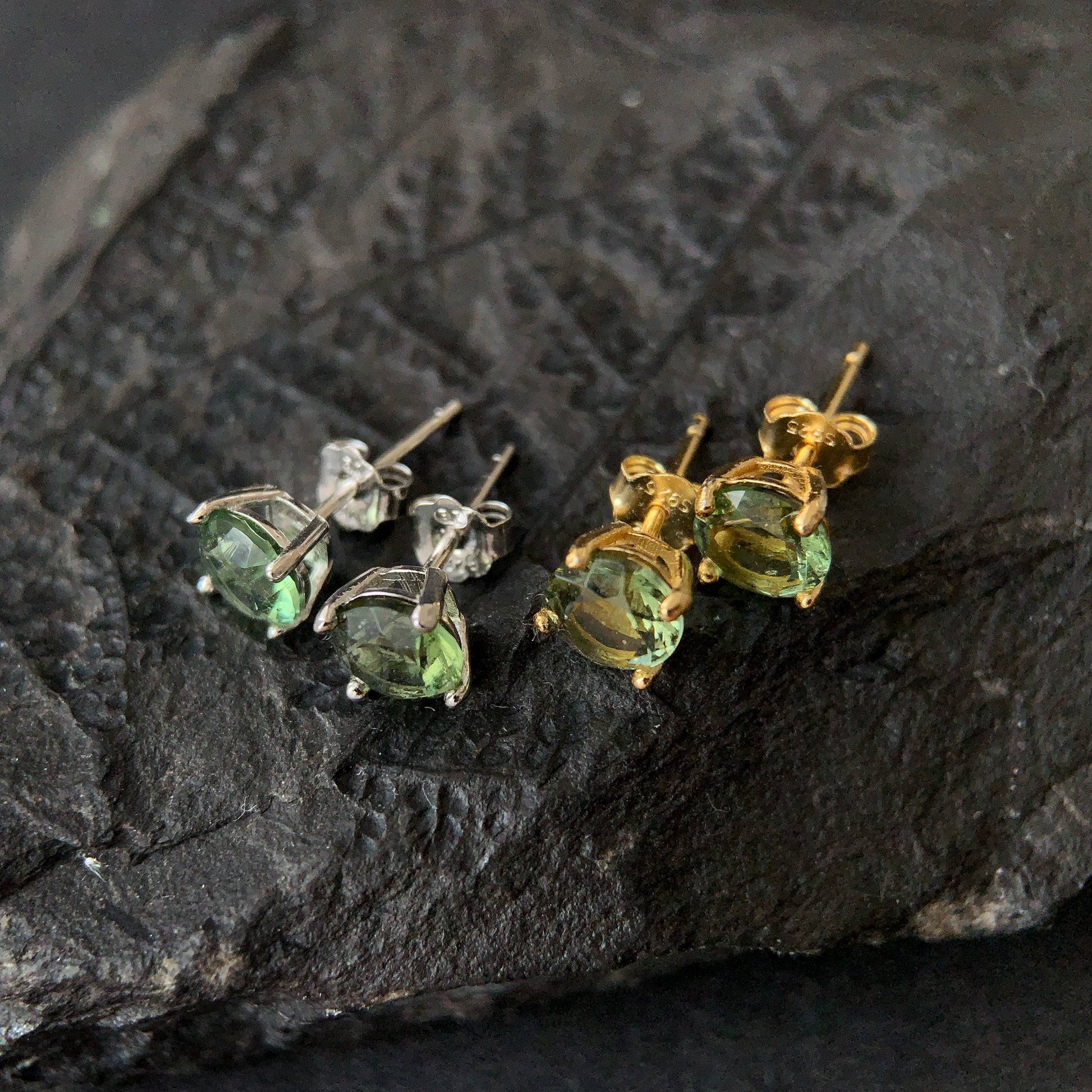 Two pairs of green gemstone stud earrings, one gold plated and one silver, on a dark stone surface.